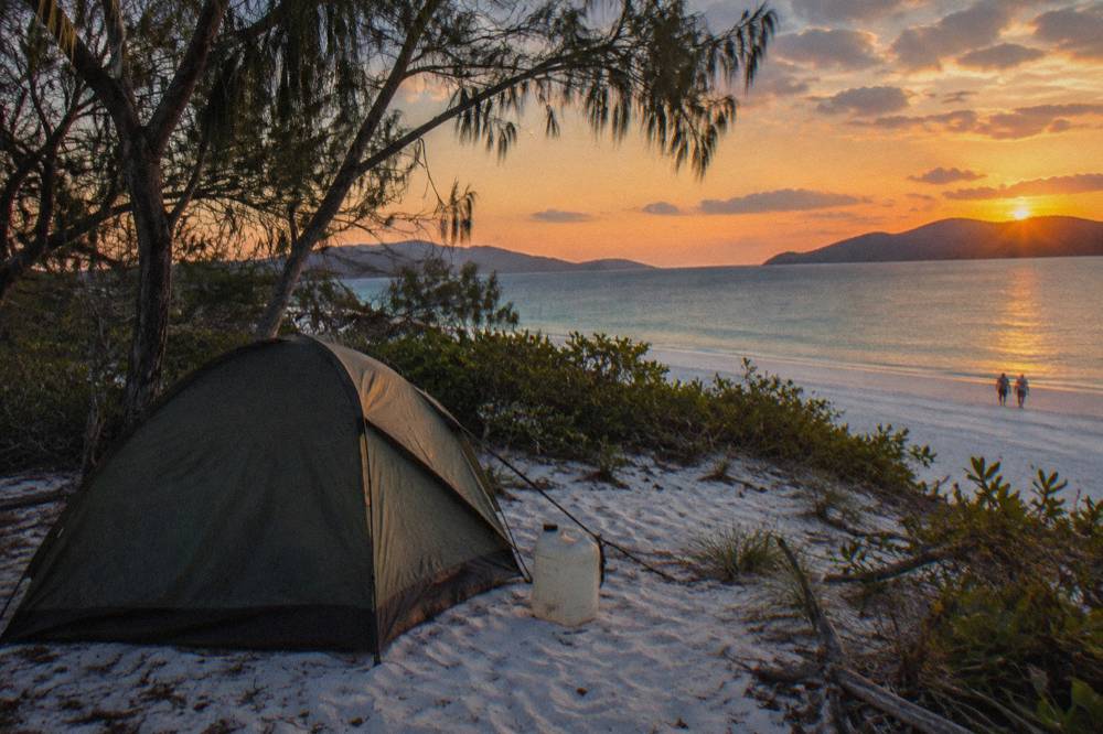 A small tent pitched behind the dune line on a white sand beach under an evening sky