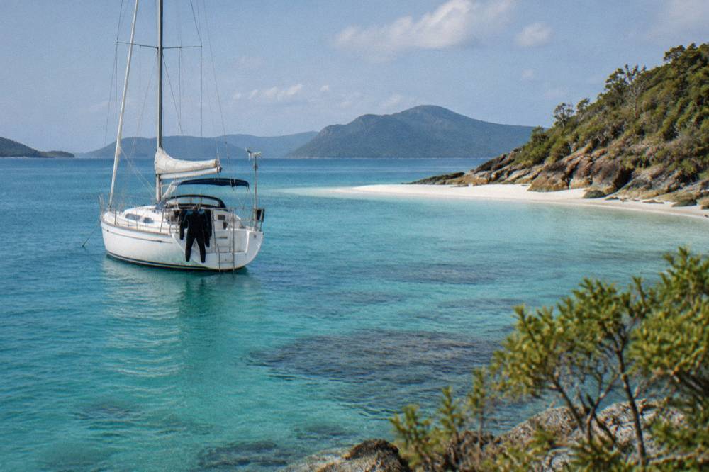 Small white sailing yacht anchored off a pale sand beach with turquoise shallows