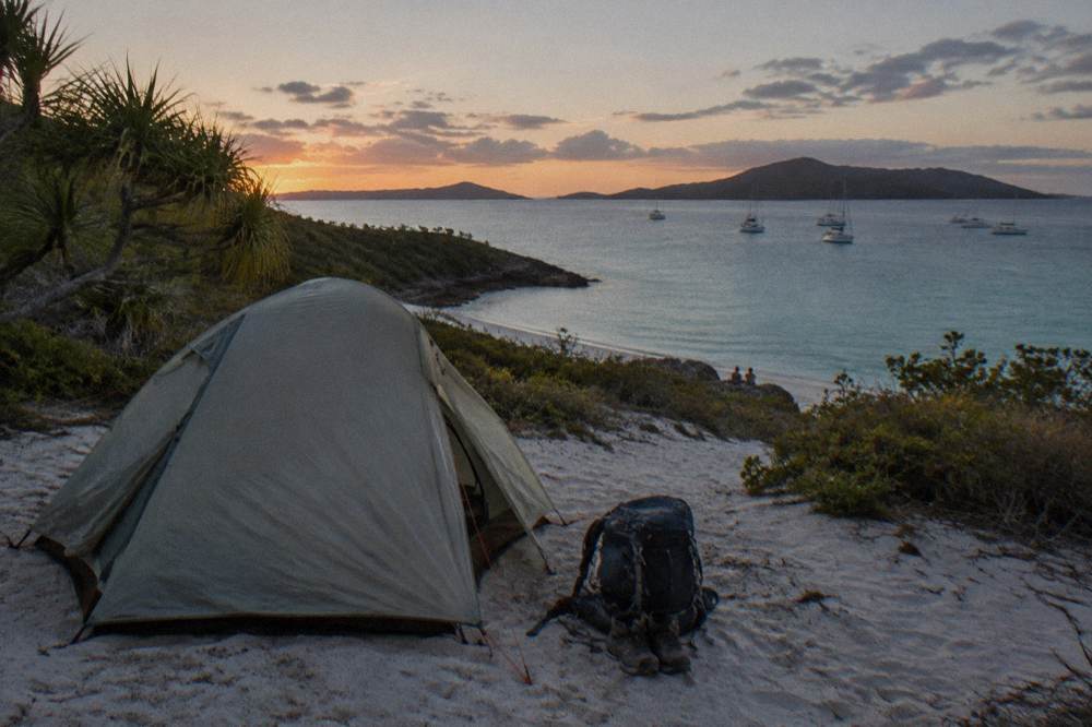 Tent on sand beside calm turquoise water at dusk with anchored boats in the distance