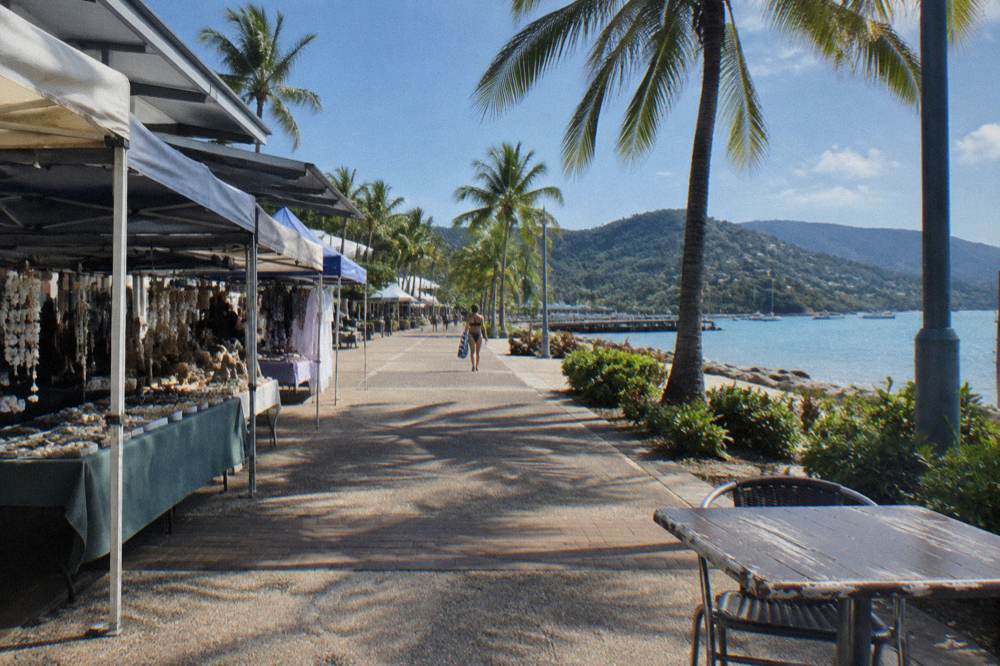 A tropical waterfront street with palm trees, cafes and market stalls near a swimming lagoon