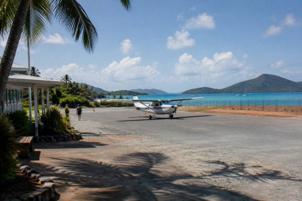 A small aircraft on a sunny runway beside tropical water under scattered clouds