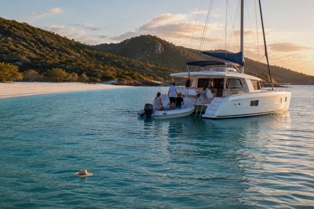 A white catamaran at anchor off a tropical island beach with passengers boarding a small tender