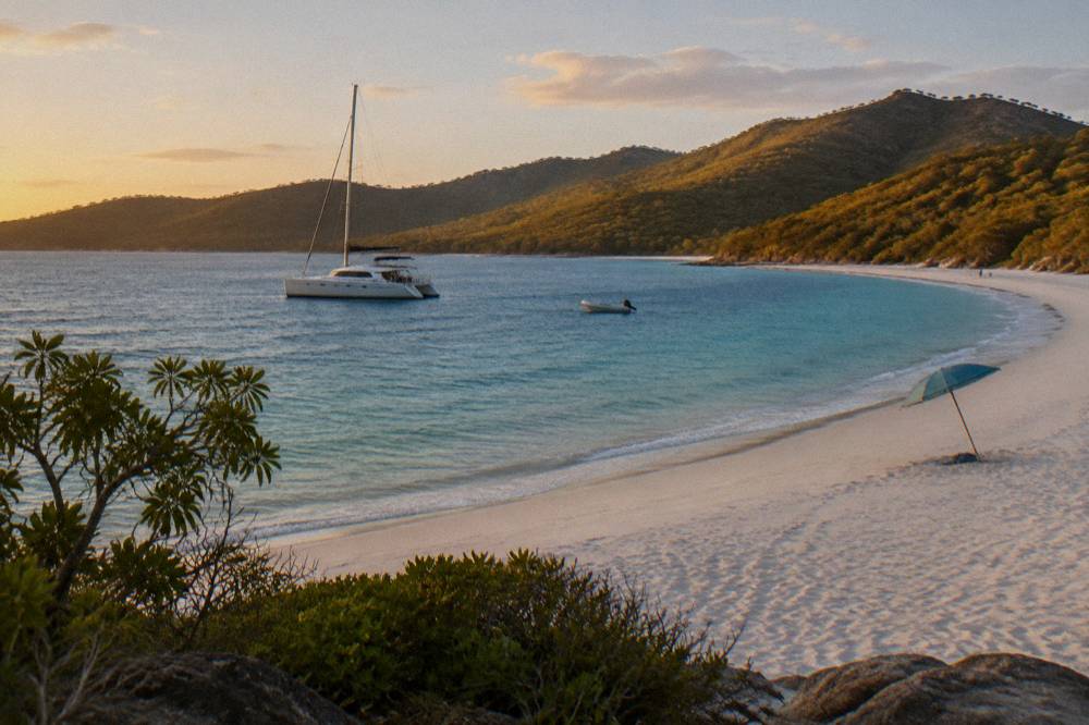Large sailing catamaran anchored off a long white beach with forested hills behind