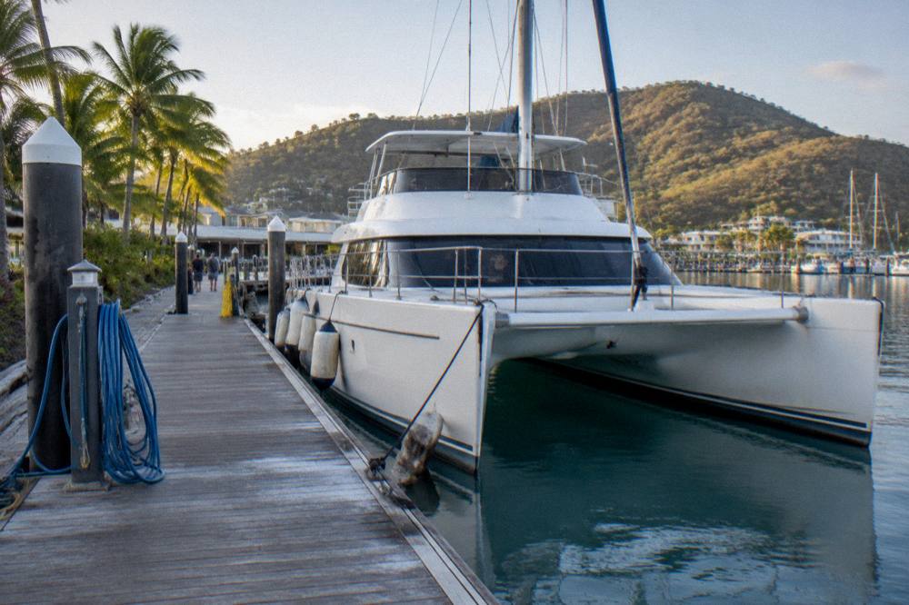 A large catamaran moored in a marina with palm trees and hills behind