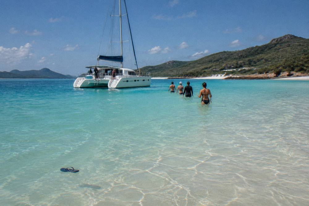 A white catamaran moored off Whitehaven Beach with passengers wading in shallow turquoise water
