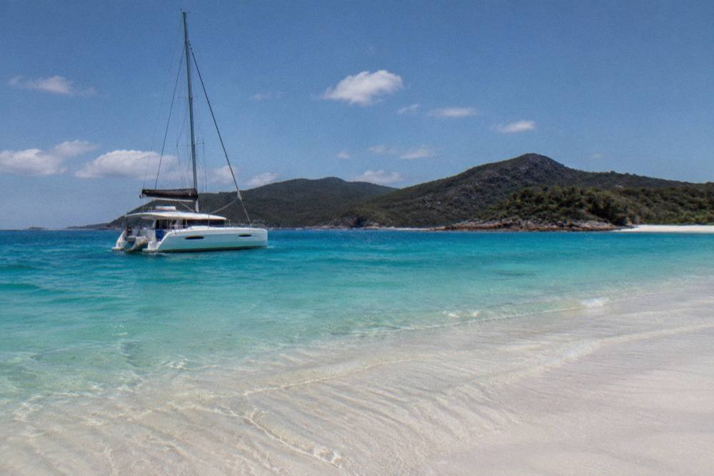 A white sailing yacht anchored off a long white sand beach with turquoise shallows