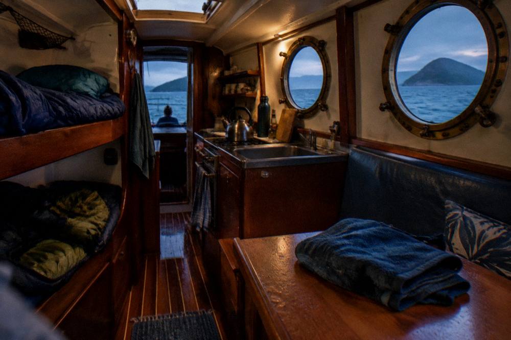 A shared yacht galley with bunks and portholes showing blue water outside