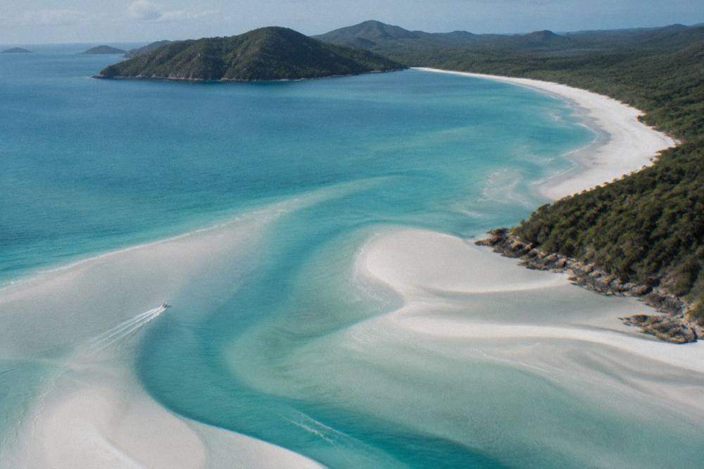 Aerial view of white sand swirls where turquoise tide meets a long white beach between green headlands