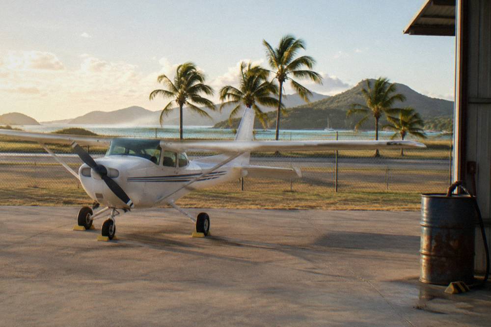 A light aircraft parked on a small regional airfield apron with palm trees at the fence line