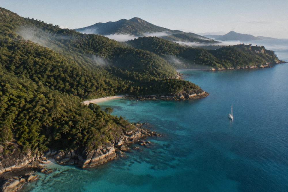 A helicopter flying low over island coastline with dark green forest meeting clear blue sea