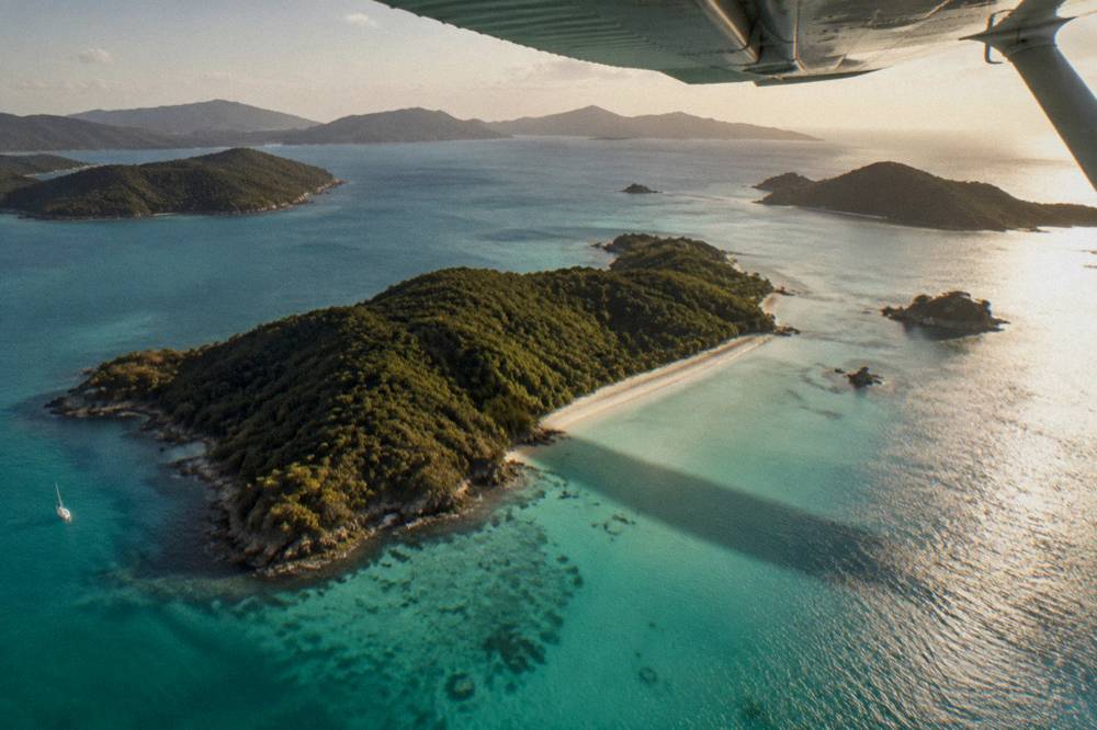 A high wing light aircraft in flight above scattered green islands and pale turquoise coral water
